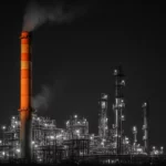 An industrial oil refinery illuminated at night with multiple chimneys and smoke.