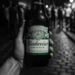 A hand holding a Budweiser beer bottle with a street scene in the background.