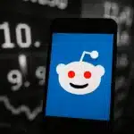 Reddit logo displayed on a smartphone screen in front of stock market graphics.