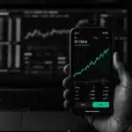 Smartphone displaying stock charts in front of a laptop with financial data.